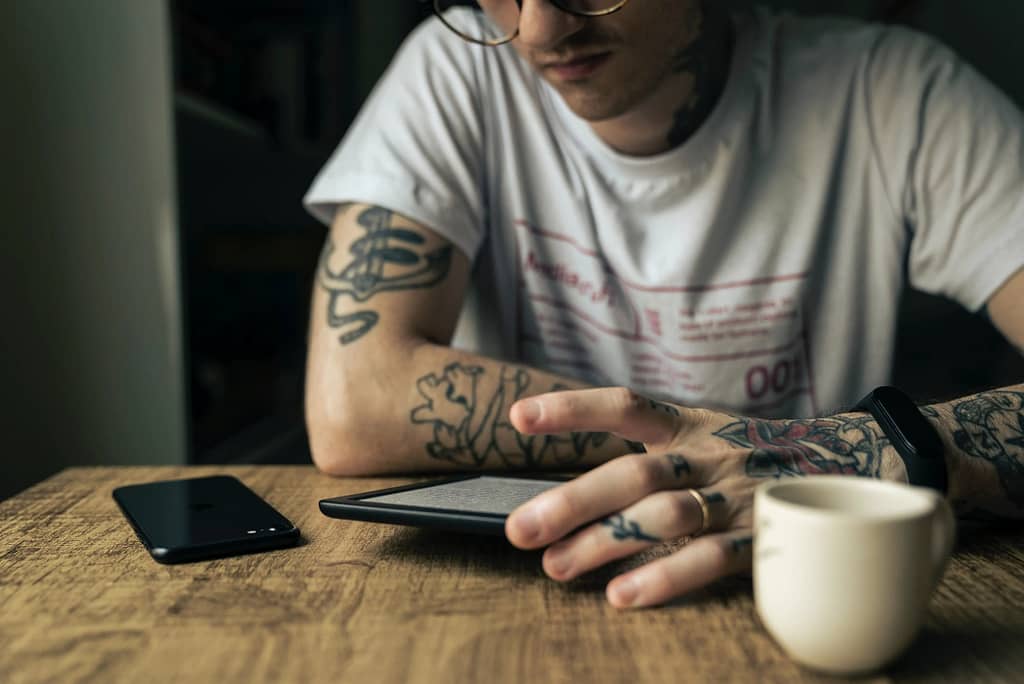 Tattooed man reading a kindle sat at a table with coffee 
